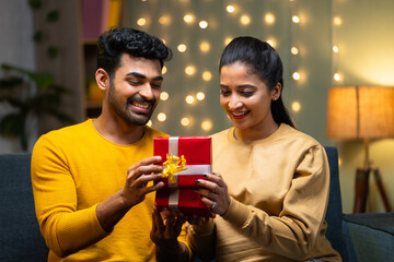 Happy Indian young man giving gift to girlfriend during wedding anniversary or birthday on sofa at home - concept of affection, dating and relationship