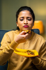 Vertical shot of young Indian girl enjoys eating tasty pizza by closing eyes at home - concept of...