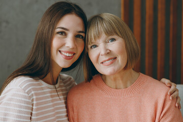 Close up two smiling happy adult women mature mom young kid wear casual clothes look camera hug sit on gray sofa couch stay at home flat rest relax spend free spare time in living room Family concept
