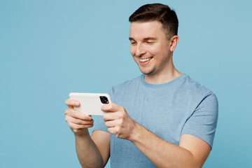 Young smiling happy fun man wear casual t-shirt use play racing app on mobile cell phone hold gadget smartphone for pc video games isolated on plain pastel light blue cyan background studio portrait.