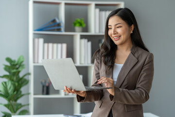 Fototapeta na wymiar Asian businesswoman smiling confidently successful entrepreneur in the financial business professional company executive Wearing suit standing holding laptop searching business information in office