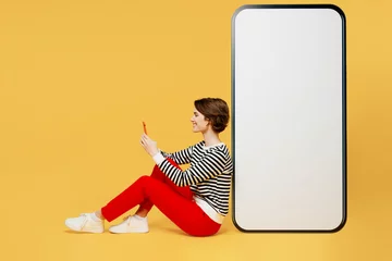 Gardinen Full body sideways happy young woman wear casual black and white shirt sit big huge blank screen mobile cell phone with workspace mockup area using smartphone isolated on plain yellow color background © ViDi Studio