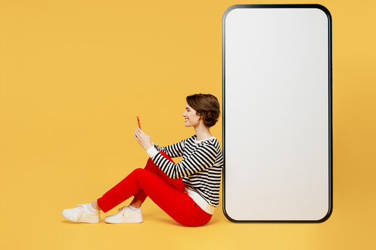 Full body sideways happy young woman wear casual black and white shirt sit big huge blank screen mobile cell phone with workspace mockup area using smartphone isolated on plain yellow color background