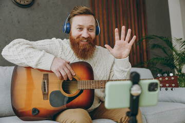 Young man wears casual clothes headphones play guitar use mobile cell phone waving hand sit on grey...