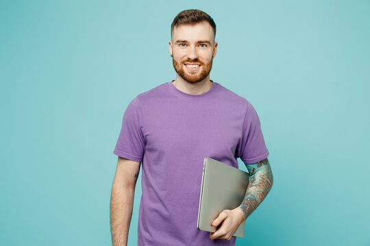 Young confident fun cheerful happy programmer smart IT man he wear purple t-shirt hold closed laptop pc computer isolated on plain pastel light blue cyan background studio portrait. Lifestyle concept.