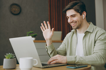 Young man wear casual clothes sit alone at table in coffee shop cafe restaurant indoors work study on laptop pc computer listen music in earphones waving hand Freelance mobile office business concept