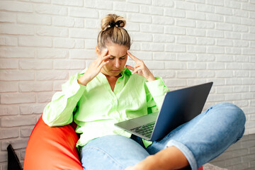 Young woman in light green shirt and jeans is uncomfortable sitting at home in armchair with laptop...