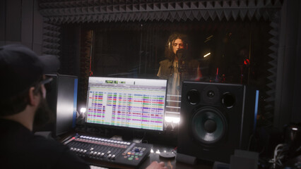 Male singer sings song in soundproof room. Audio engineer, producer uses professional DAW software...