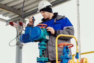Fototapeta premium An engineer with a screwdriver repairs equipment at a gas station. Male engineer in helmet and winter clothing doing maintenance on industrial gas equipment outside..