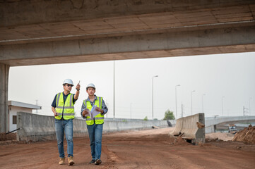 Fototapeta premium Asian Two civil engineer specialists examining trades at road construction site, industrial building construction site. Concrete skyscraper of road construction
