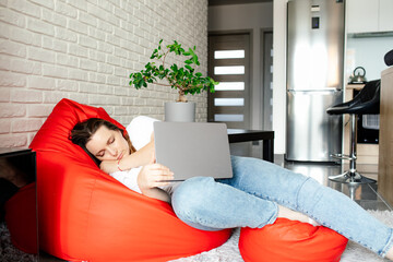 Young woman in white T-shirt and jeans fell asleep with laptop on her lap, sitting at home in...