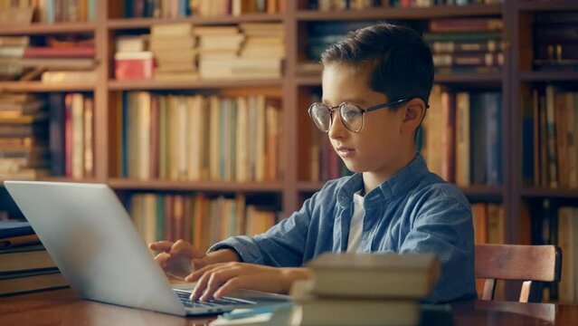 Diligent schoolboy in glasses studying online on laptop at library, typing essay