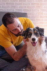 Young man sitting on the sofa petting his female dog (Australian Shepherd). The dog looks at the camera.