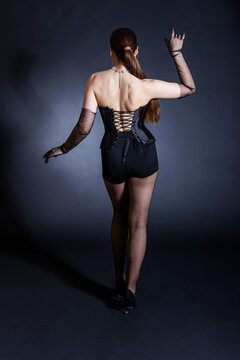 A seductive young woman in a black corset poses elegantly against a dark background, standing with her back to the viewer.