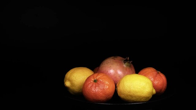 Female hand continuously, piece by piece picking up fresh organic lemons, pomegranates and mandarins from a black plate. Beautiful citrus fruits. Isolated black background with copy space. 4K