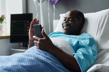 African American man smiling while talking online with his relatives on smartphone, he lying on bed...
