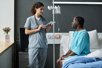 Young nurse asking about health condition to patient and typing information in digital tablet...