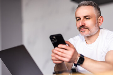 Old man sit in kitchen near laptop holding smartphone device chat remotely, browse webpage, use new phone app enjoy usage of modern wireless tech, make business from home distantly concept