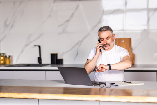 Middle age man talking on the smartphone, look at watch and using laptop at home