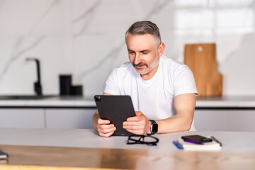 Middle-aged man looking at camera and checking messages on digital tablet while having breakfast on kitchen counter at home