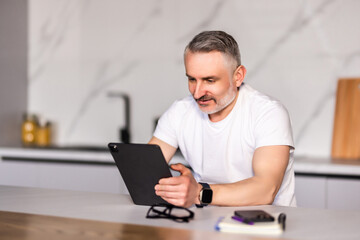 Middle-aged man looking at camera and checking messages on digital tablet while having breakfast on kitchen counter at home