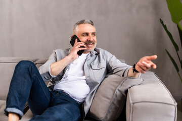 Cheerful handsome middle-aged bearded man enjoying weekend at home alone having conversation on mobile phone with friends or lover, sitting on sofa, looking at copy space