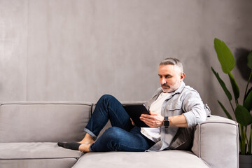 Portrait of a serious handsome bearded guy, young middle aged man is using his tablet laptop, relaxing, lying at home on sofa couch, looking at screen, reading electronic book