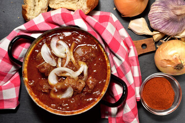 Pork stew in the old enameled saucepan on a checkered red tablecloth. Onion, garlic and pepper spice. View from top.