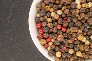 A mixture of four peppercorns on a white saucer, macro, on slate stone, top view.