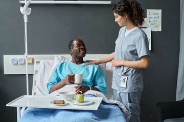 Young nurse in uniform serving lunch to senior patient while he sitting on bed in ward