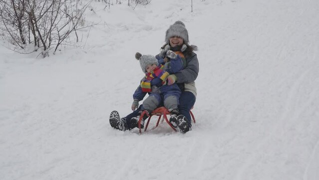 Cheerful mom and son on sleigh enjoy winter by sliding on snow slope