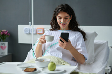Young girl having lunch and watching video on smartphone while sitting on bed in ward