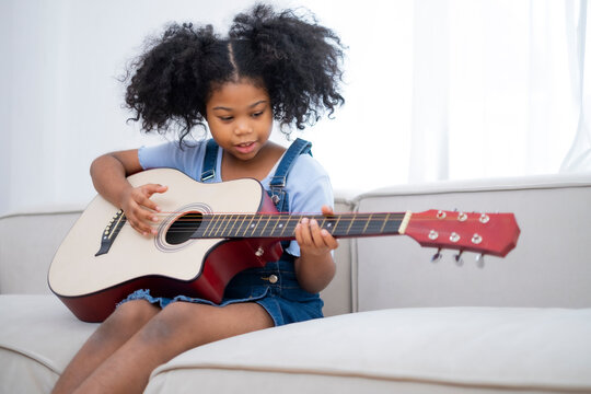 African American little girl enjoy play guitar at home, student practice music instrument sitting on coach. Female Black child play guitar on sofa.