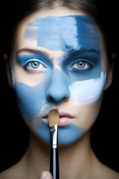 Generative AI illustration of young female model with blue eyes and face painted in blue and white colors looking at camera while holding makeup brush on lip against black background during beauty mak