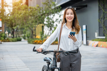 Naklejka na ściany i meble Portrait of beautiful smile business woman commute her bicycle outdoor using smartphone at urban, bike go to work office, Asian businesswoman standing on city street with bicycle holding mobile phone