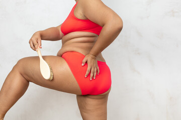 Overweight woman with fat folds standing with back do dry thigh massage with brush, free copy space, white background. Large woman in red underwear get rid of cellulite. Plus size people, weight loss.