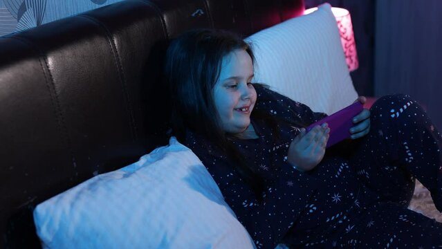 Portrait of happy preteen girl with loose dark hair wearing pajama with stars, lying on bed on white pillows at night, holding smartphone, playing games, tapping screen having fun. Technology, gaming.