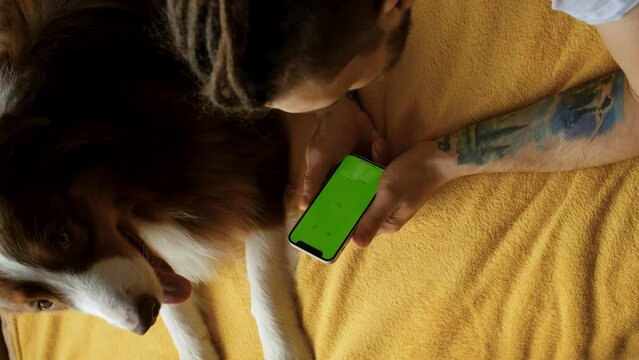 Young man lying on yellow blanket inside next to pet dog Australian shepherd and resting together. Vertical 4k footage. Buying animal food. Using Mobile with green screen Chroma Key Device.