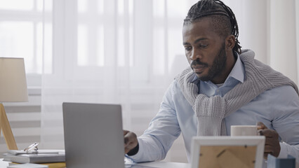 African American man programmer typing on laptop, drinking coffee at home, work from home