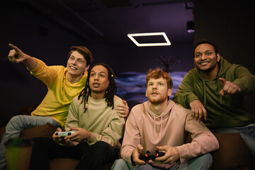 KYIV, UKRAINE - FEBRUARY 13, 2023: Smiling interracial men pointing with fingers while friends playing video game in gaming club.
