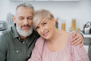 happy bearded man with closed eyes embracing wife smiling at camera in kitchen.