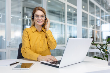 Portrait businesswoman, office worker looking at camera and smiling, using headset and laptop for...
