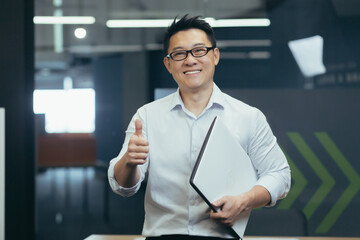Portrait of a successful young Asian male programmer standing in a modern office with a notebook in...