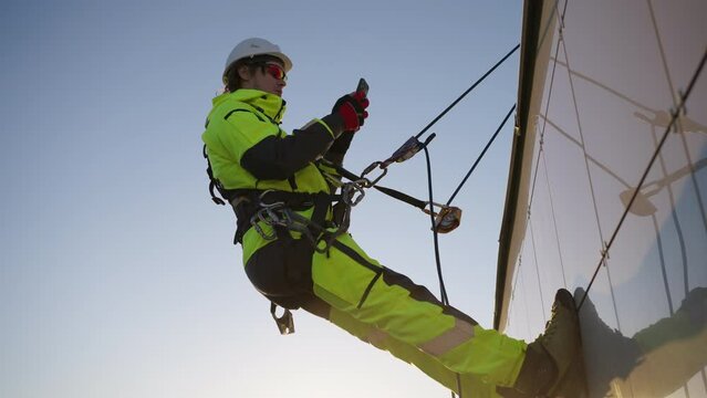 Man industrial climber in equipment and a helmet is tied with a rope to a building with a phone in his hands looking at the sunset