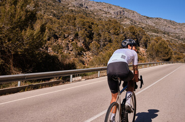 Fit man cyclist wearing white cycling jersey and helmet training on road bicycle on empty mountain...