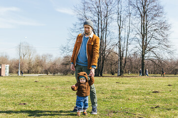 a young father with a little son in a bear-shaped hoodie stand in the park