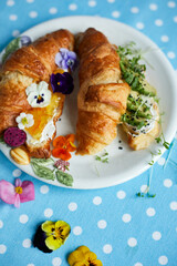 Side view of a croissant with orange jam and cream cheese and with avocado, microgreens on a white plate, pansies, fruits. Healthy breakfast.