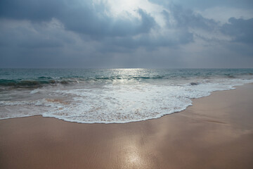 A gorgeous view of a sandy ocean beach with turquoise waves under a dramatic cloudy sky with sunlight through it.