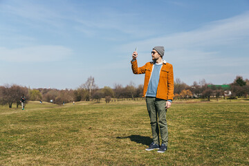 a man in a hat and an ocher corduroy jacket stands in a field in a park with a phone in his hands...