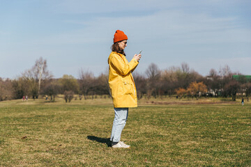 a girl in a yellow jacket and a red hat with glasses stands sideways in full growth in the park and looks at the smartphone screen